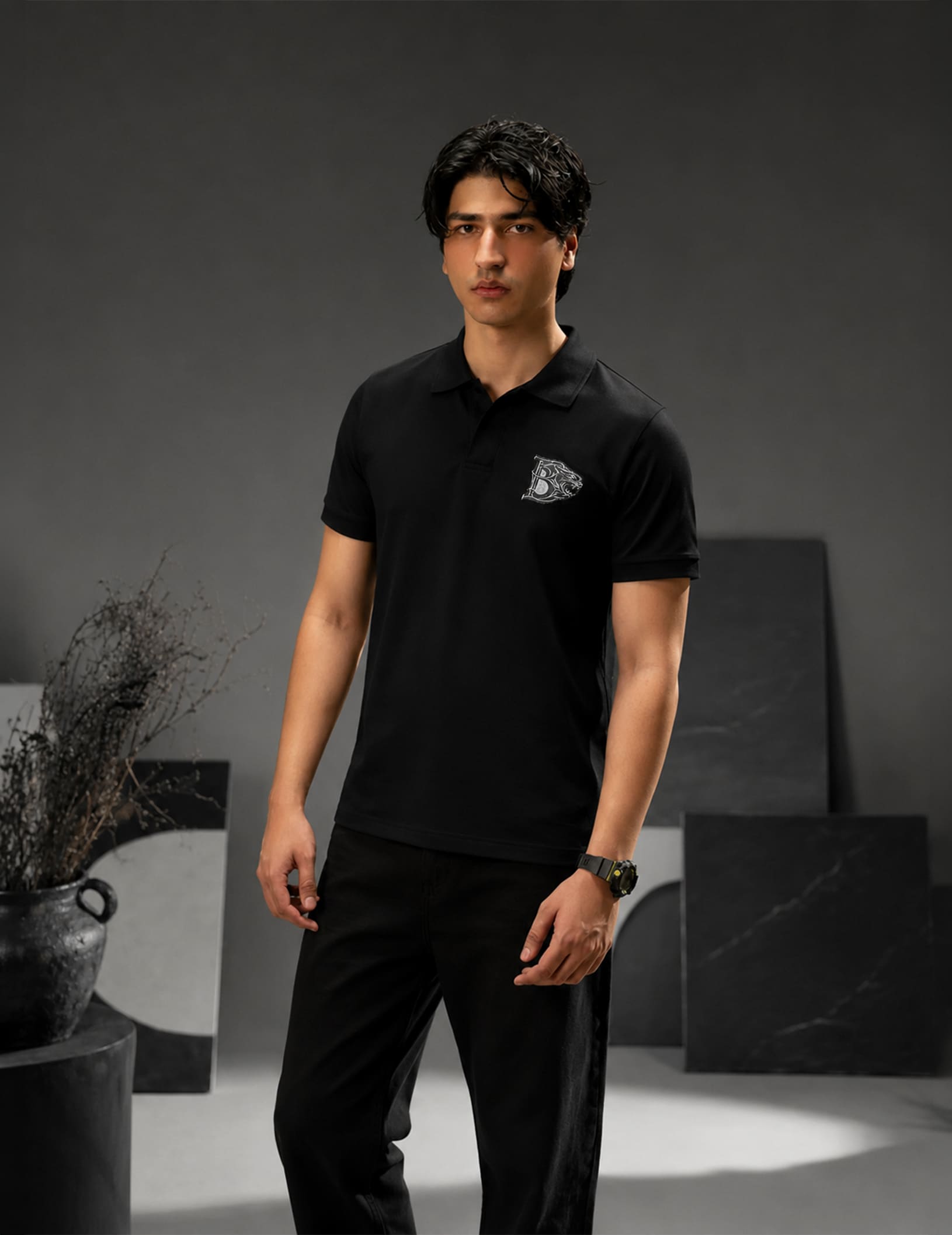 Male model wearing the BLACOIR Crest V-Collar Tee featuring a tonal crest logo embroidery, styled in a sophisticated luxury editorial environment with black-on-black aesthetics.