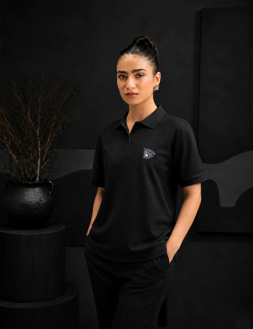 Female model wearing the BLACOIR Crest V-Collar Tee with a tonal silver crest emblem, paired with black trousers in a premium minimalist fashion studio.