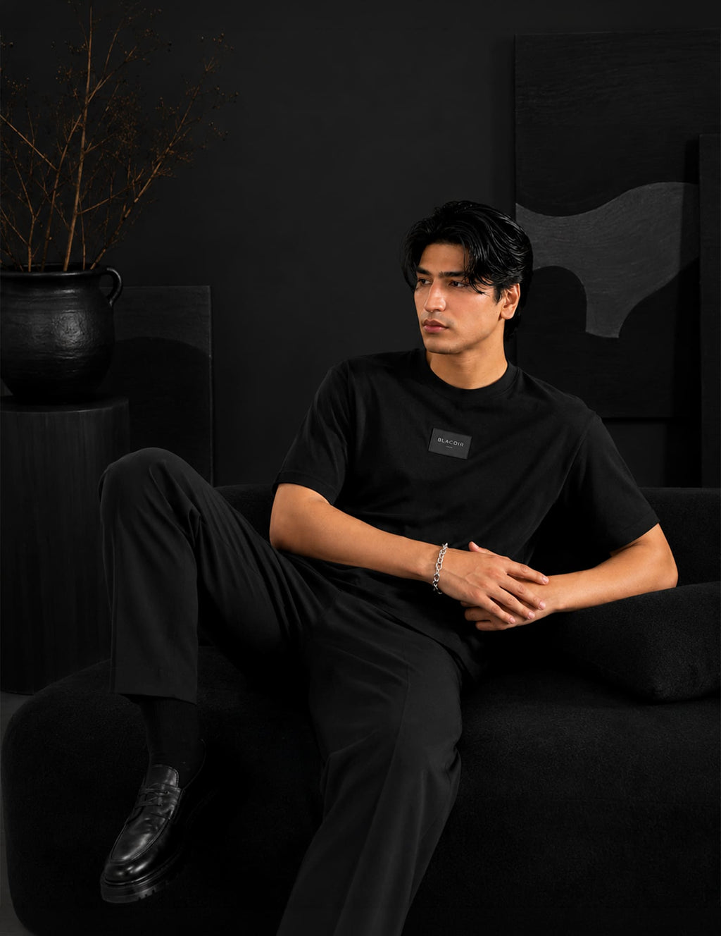 Male model reclining on a black sofa wearing the BLACOIR Essential Black Tee with tonal logo patch, black tailored trousers, and luxury minimalist styling in a dark studio setting.