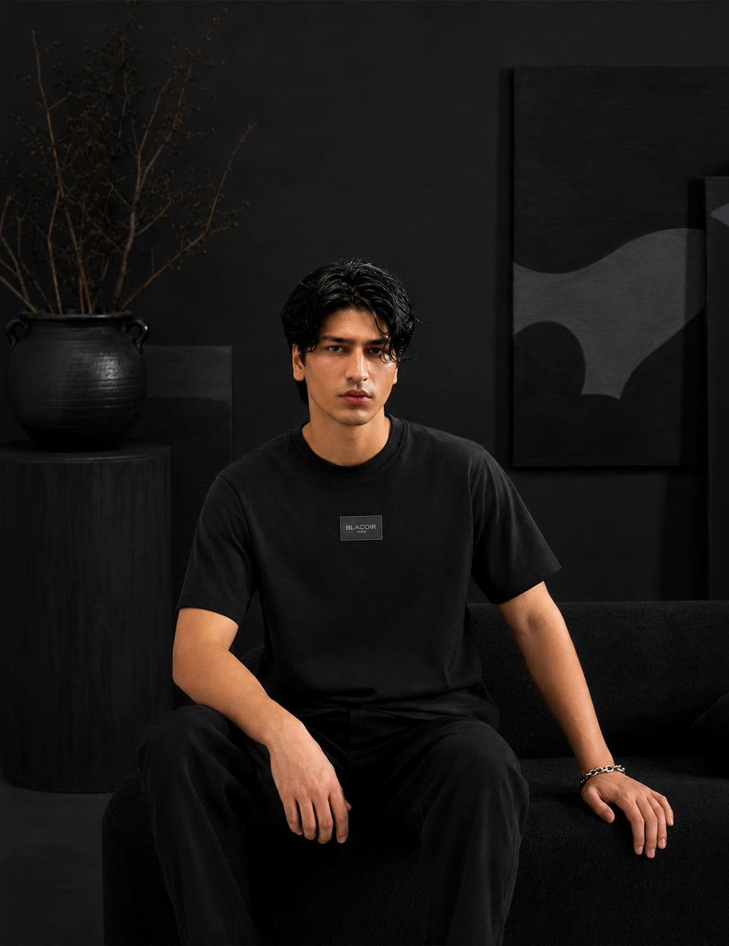 Male model seated in a luxury black interior wearing the BLACOIR Essential Black Tee with tonal chest logo patch, styled with black trousers for a sophisticated monochromatic look.