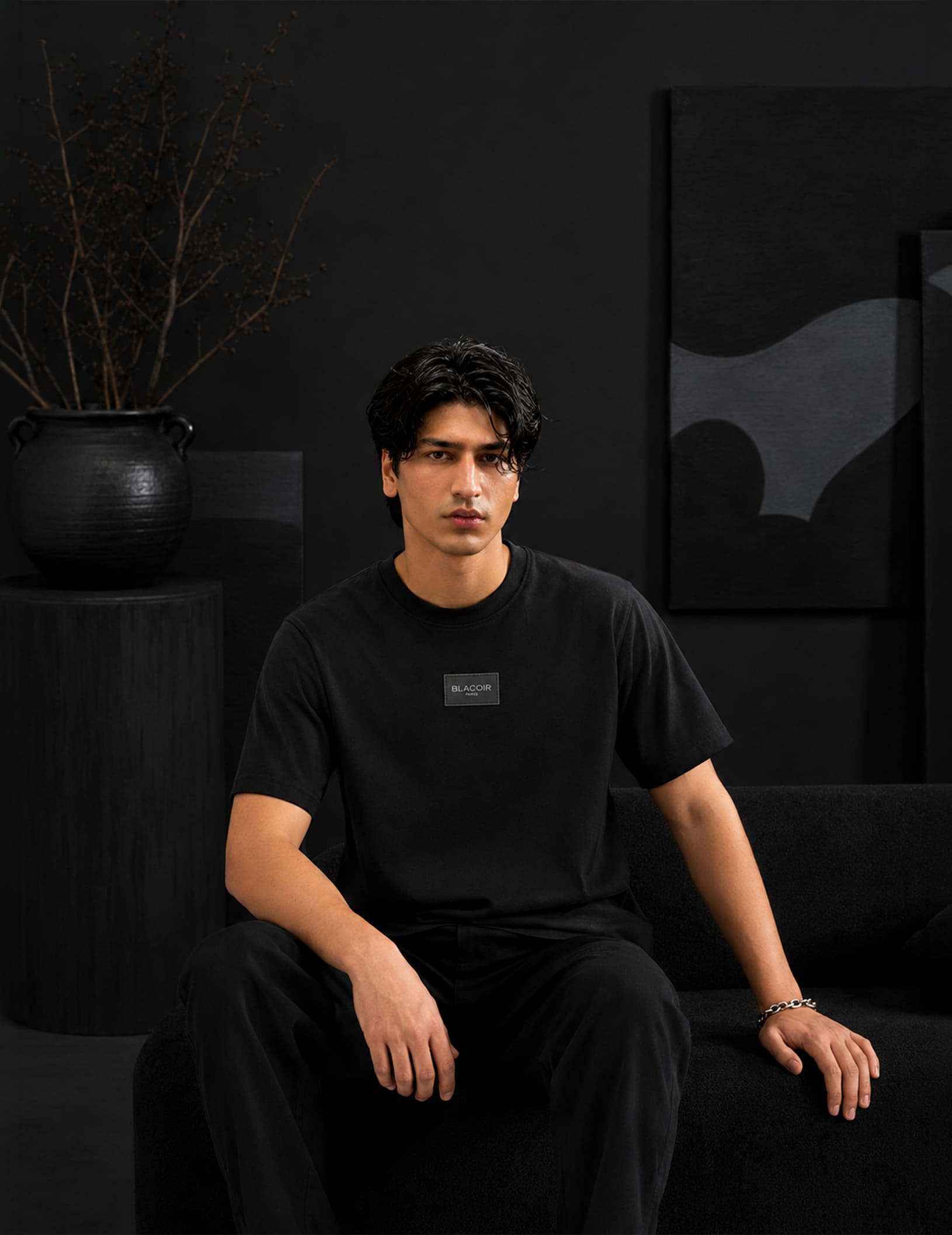 Male model seated in a luxury black interior wearing the BLACOIR Essential Black Tee with tonal chest logo patch, styled with black trousers for a sophisticated monochromatic look.