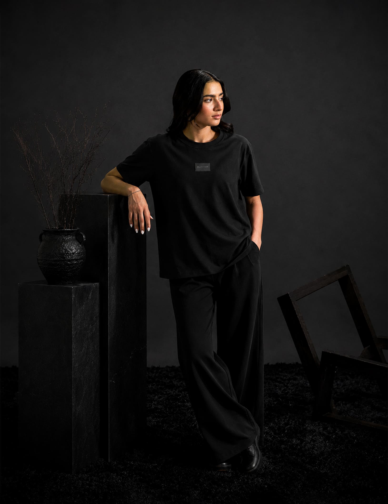 Female model wearing the BLACOIR Essential Black Tee with tonal BLACOIR Paris chest patch, styled with wide-leg black trousers in a premium monochromatic fashion studio.
