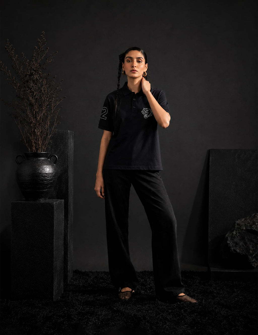 Female model seated in a luxury black armchair wearing the BLACOIR Panther Polo featuring a tonal panther chest graphic and sleeve number detail, styled in an elegant monochromatic setting.
