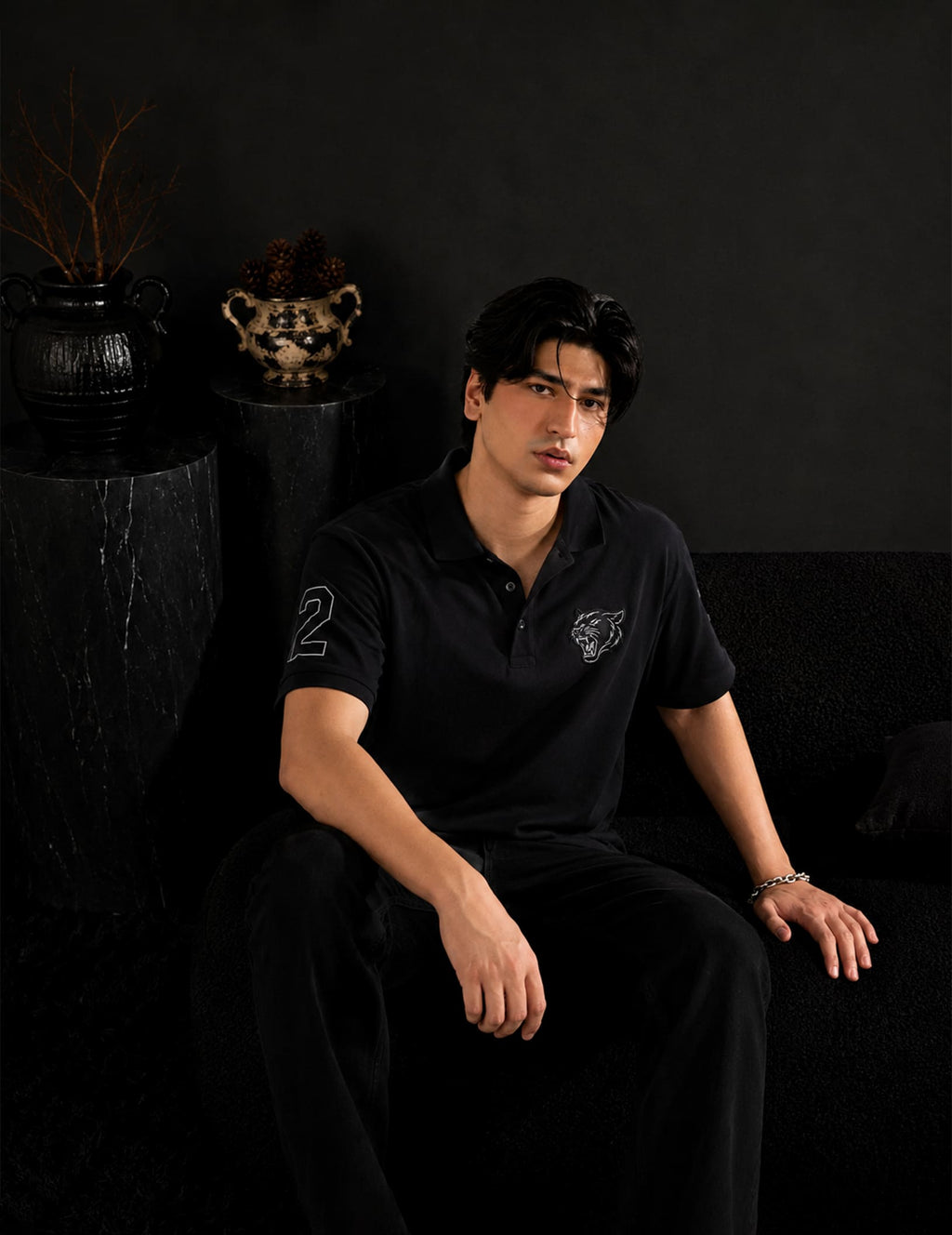 Male model seated in a luxury black studio setting wearing the BLACOIR Panther Polo in black with a tonal panther chest emblem, sleeve number detail, and black trousers.