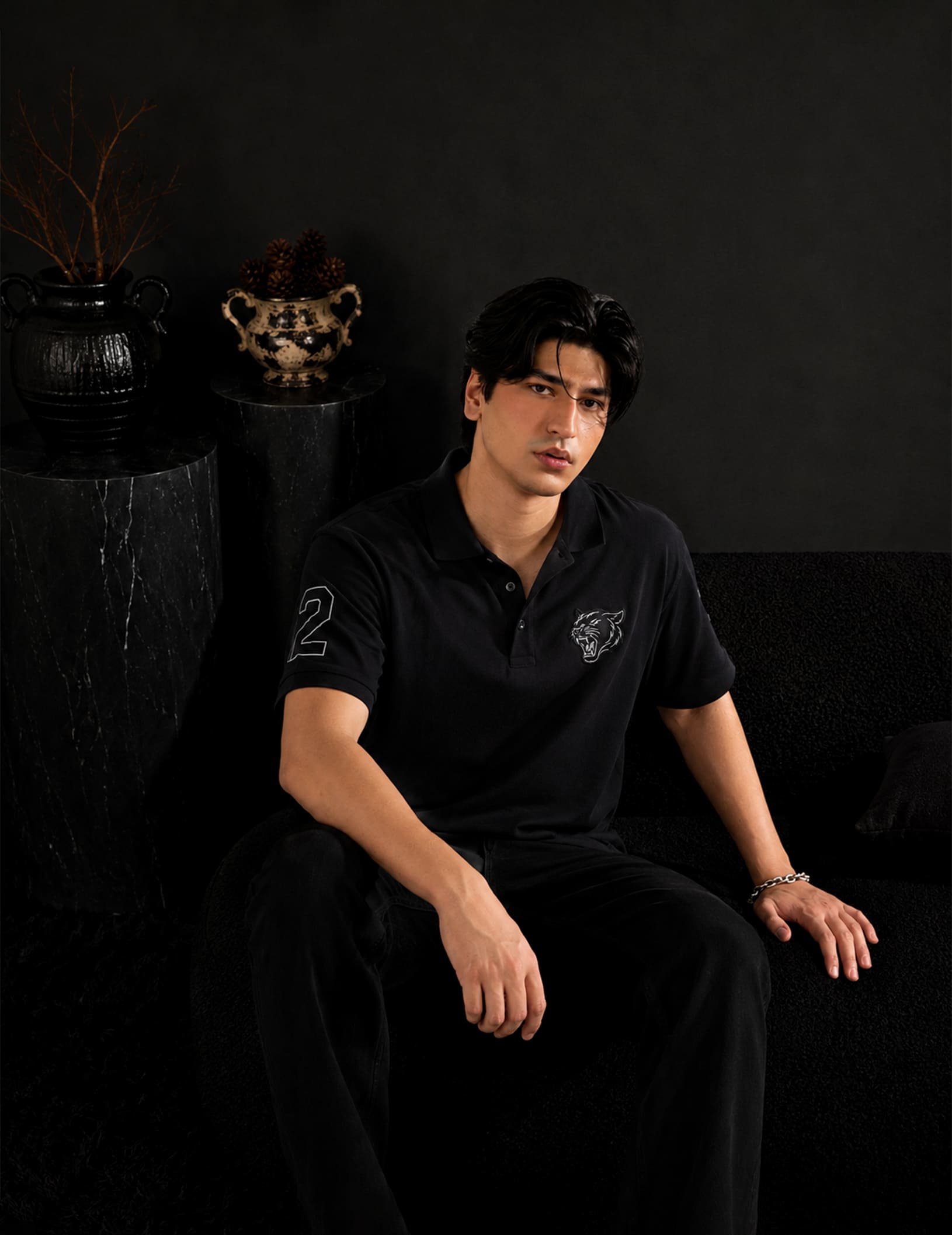 Male model seated in a luxury black studio setting wearing the BLACOIR Panther Polo in black with a tonal panther chest emblem, sleeve number detail, and black trousers.
