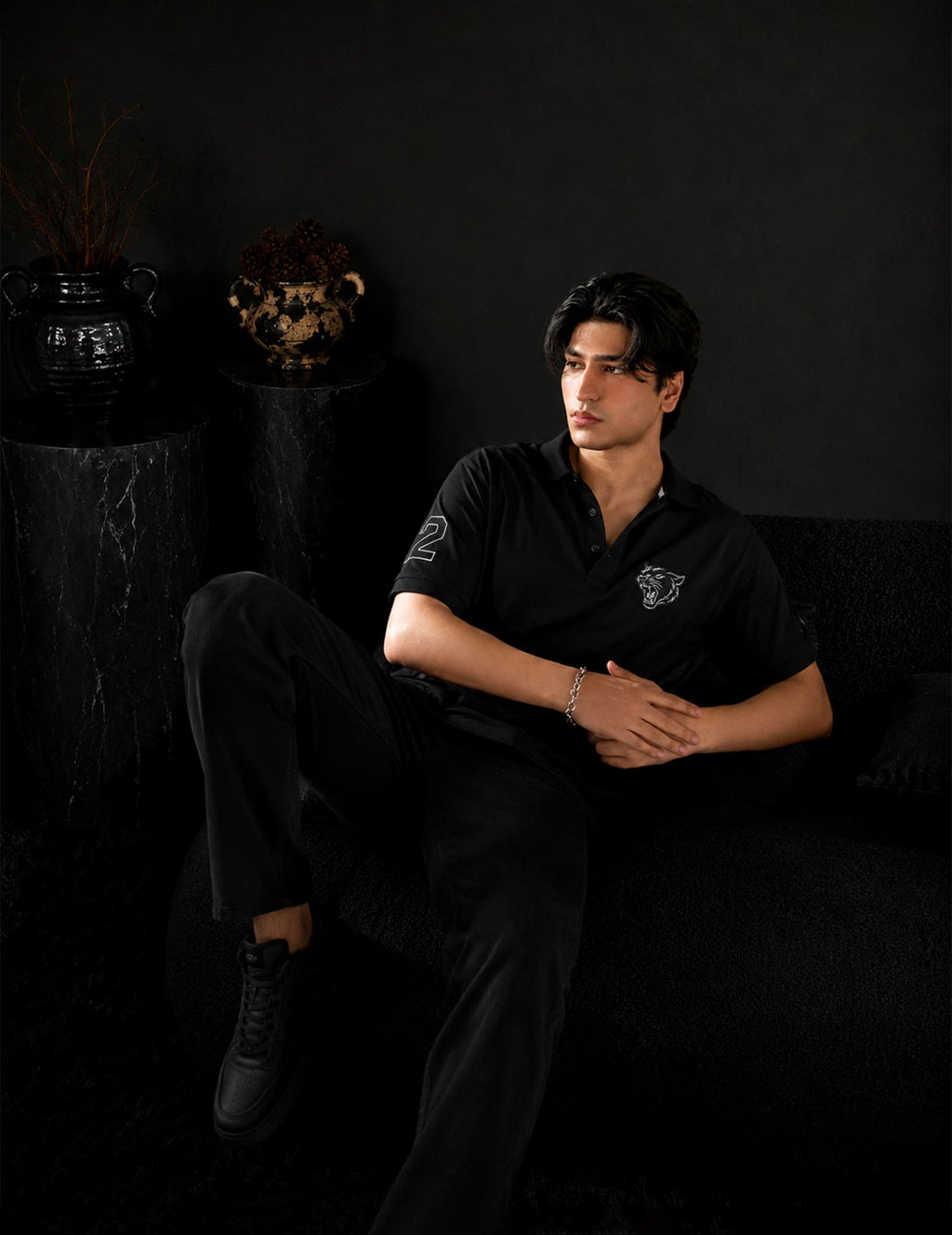 Male model seated in a luxury black studio setting wearing the BLACOIR Panther Polo in black with a tonal panther chest emblem, sleeve number detail, and black trousers.