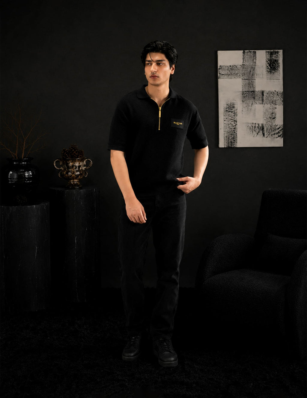 Male model wearing the BLACOIR Paris black zip polo with gold zipper detail and black denim trousers in a luxury black interior setting. Premium modern menswear from The Core Edit collection.