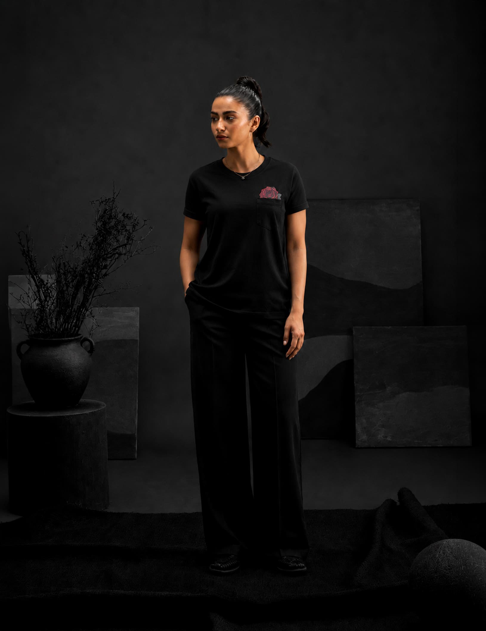 Female model wearing the BLACOIR Rosemark Pocket Tee featuring a red rose pocket artwork, paired with black wide-leg trousers in a premium monochromatic fashion studio.