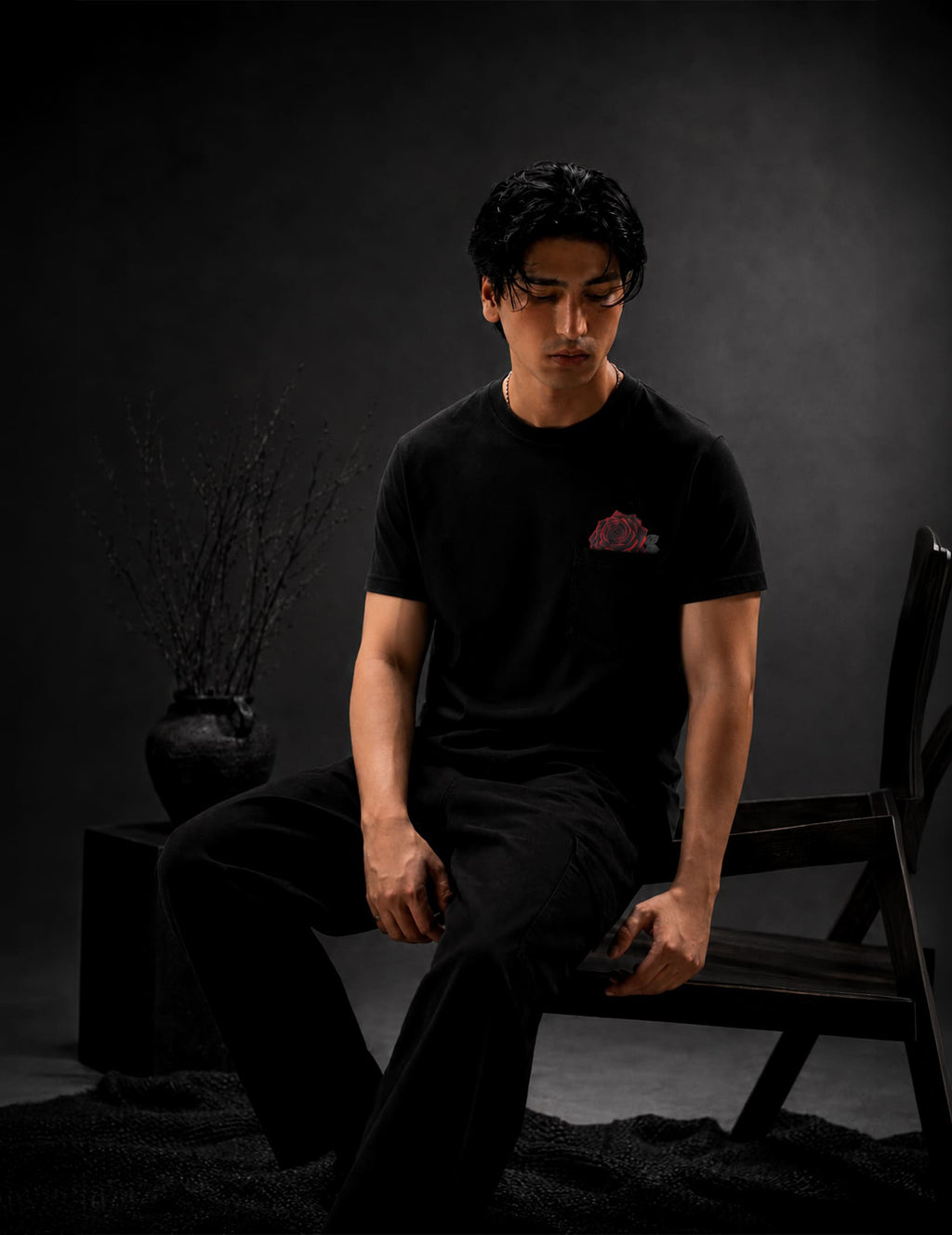 Male model wearing the BLACOIR Rosemark Pocket Tee with a red rose emerging from the chest pocket, photographed in a sophisticated black-on-black luxury environment.