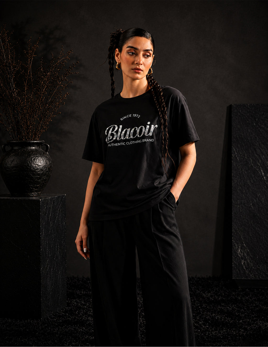 Female model wearing the BLACOIR The Glint Script Tee featuring a reflective silver glitter script logo on a premium black t-shirt, photographed in a sophisticated luxury studio environment.