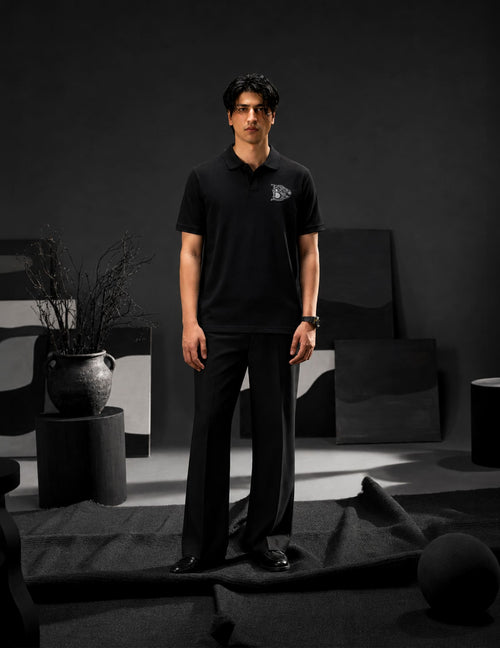 Male model wearing the BLACOIR Crest V-Collar Tee in black with a tonal crest chest emblem, styled with black tailored trousers in a modern luxury monochromatic studio.