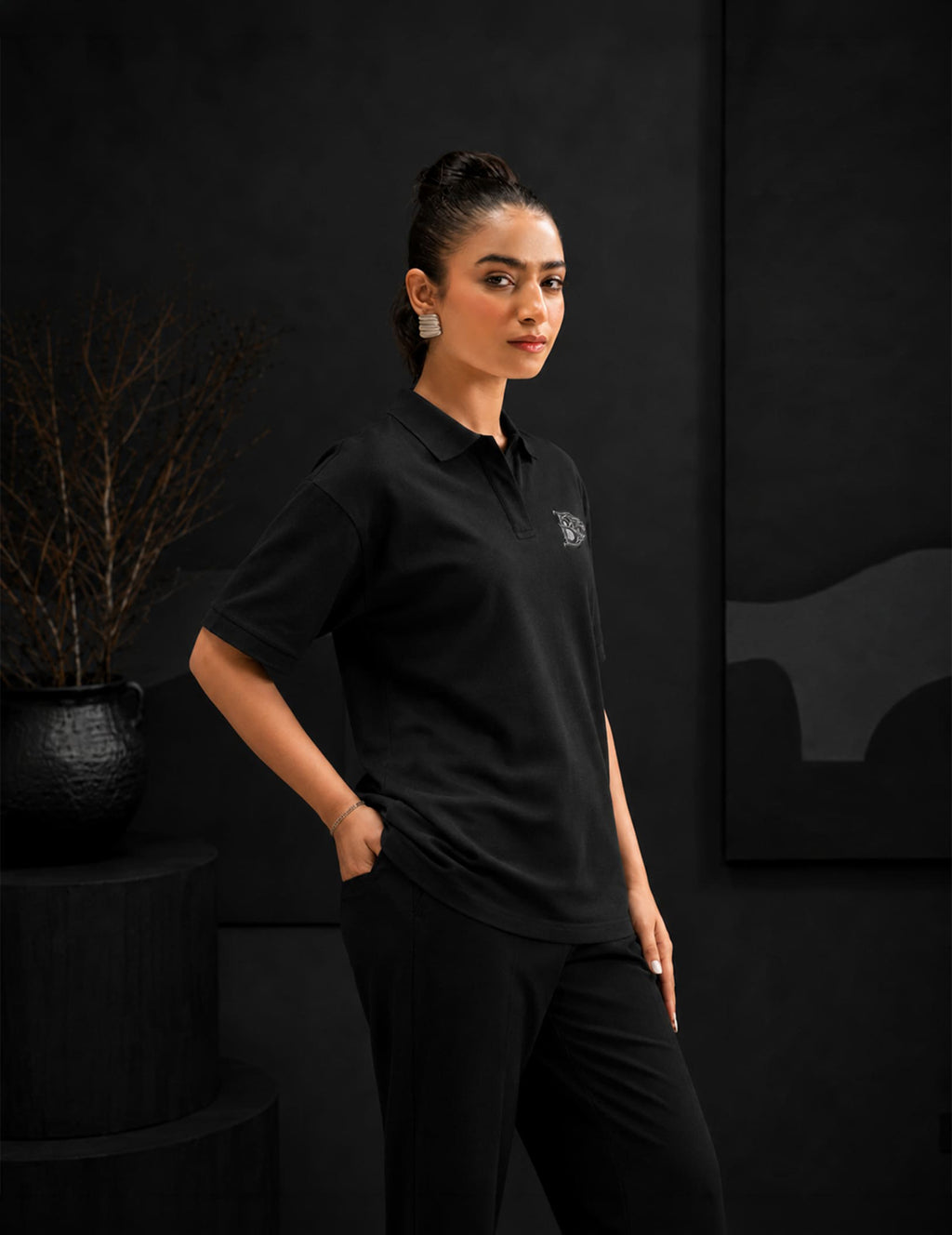 Female model in profile wearing the BLACOIR Crest V-Collar Tee featuring a luxury crest chest emblem, photographed in an elegant monochromatic editorial setting.