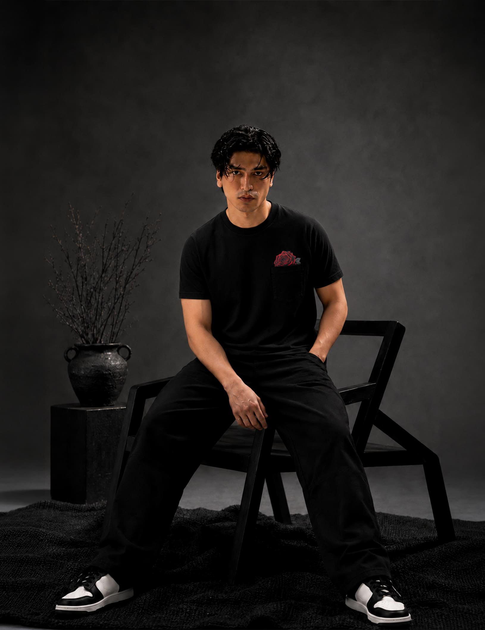 Male model wearing the BLACOIR Rosemark Pocket Tee in black, featuring a signature red rose pocket graphic, styled with black trousers in a luxury editorial studio setting.