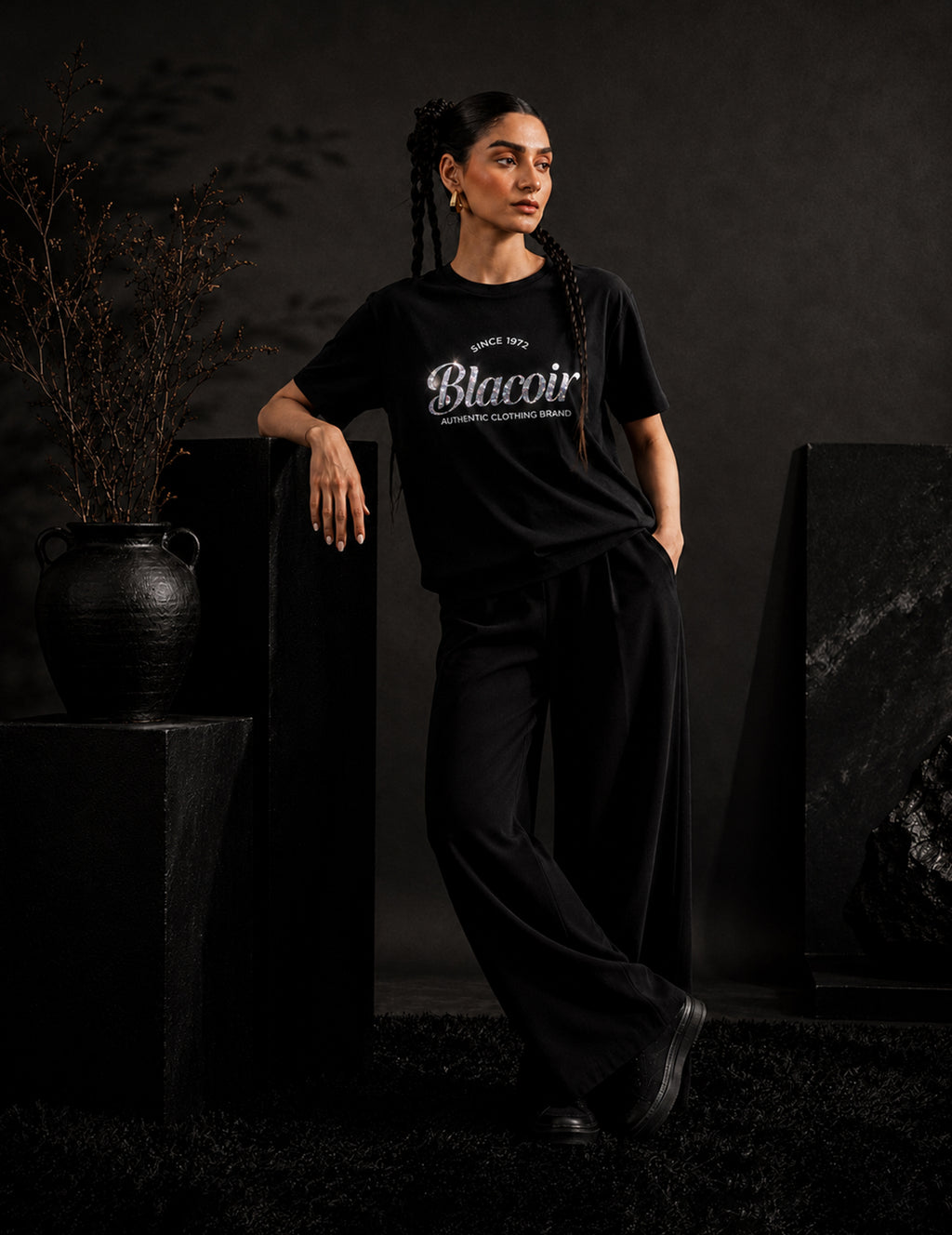 Female model wearing the BLACOIR The Glint Script Tee with metallic glitter script branding, styled with black wide-leg trousers in a luxury monochromatic editorial setting.